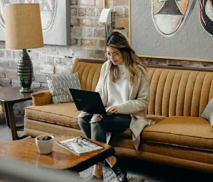 A woman sitting on a couch, using a laptop to manage a multi-channel marketing automation tool.